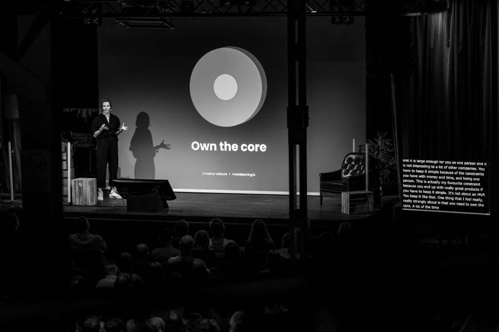 A black and white photo of Christine Vallaure on stage at beyond tellerrand in Berlin. The slide in her back shows a large circle and under the circle it says “Own the core”