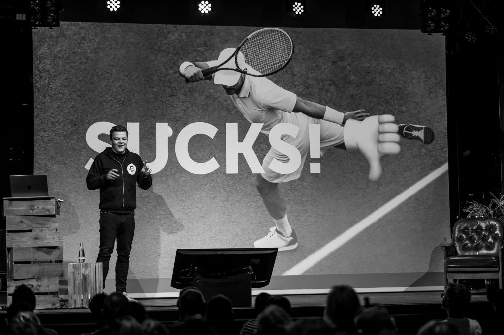 Manuel Matuzović presenting on stage at beyond tellerrand 2024. He has a smile in his face. The slide on the canvas in his back shows a tennis player from above and big capital letters say “SUCKS!”.