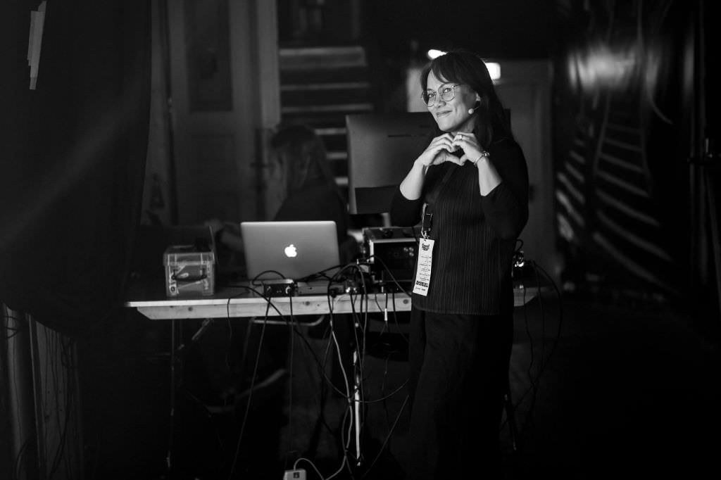 A black and white photo of Sheryl Cababa backstage at beyond tellerrand in Berlin. She looks into the camera smiling and forming a heart with her hands.