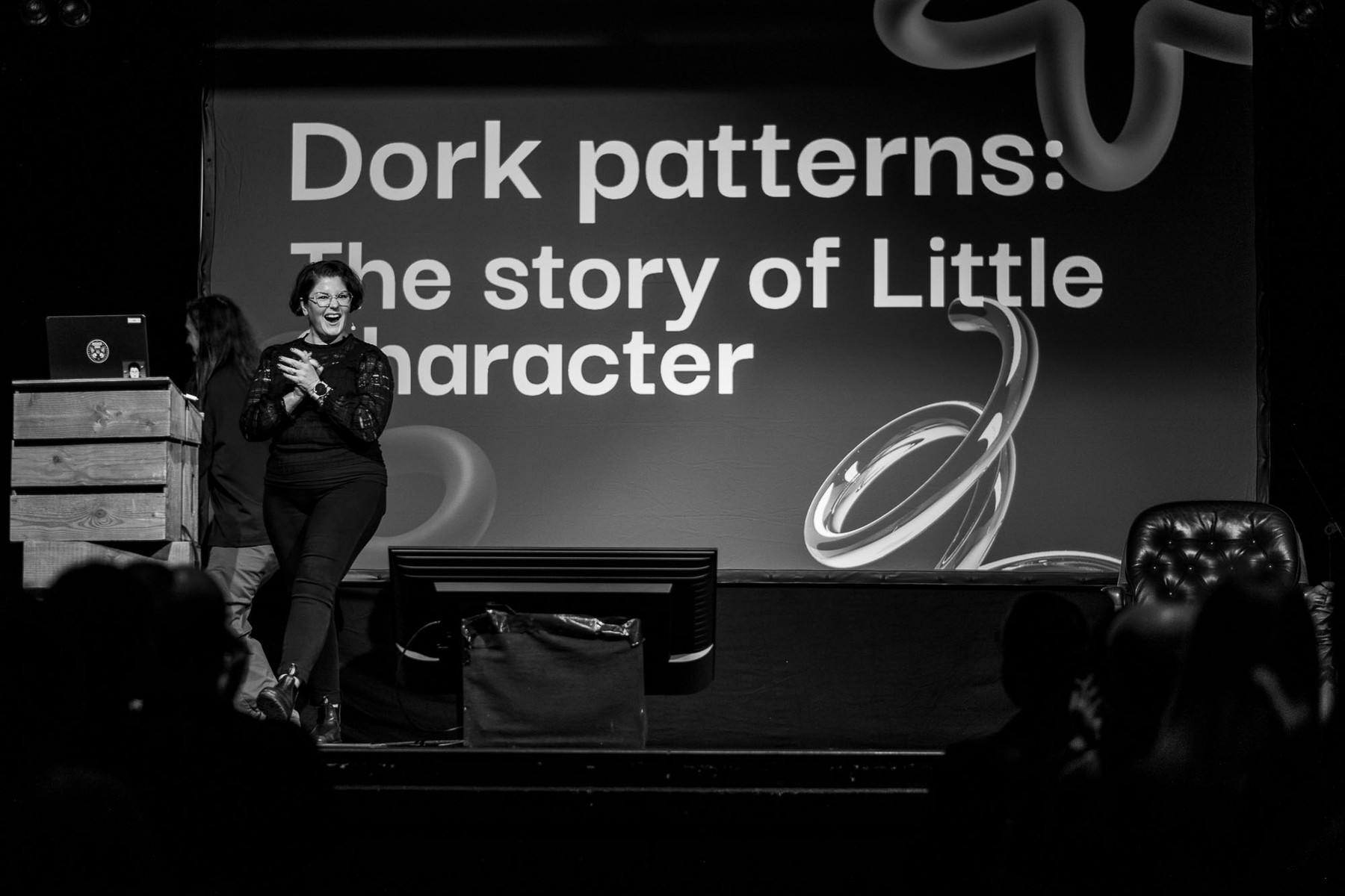 A black and white photo showing Astrid Bin on stage at beyond tellerrand right next to the speaker podium. She is laughing full of excitement and rubbing her hands against each other as a pose to get started with her talk and being full of energy.