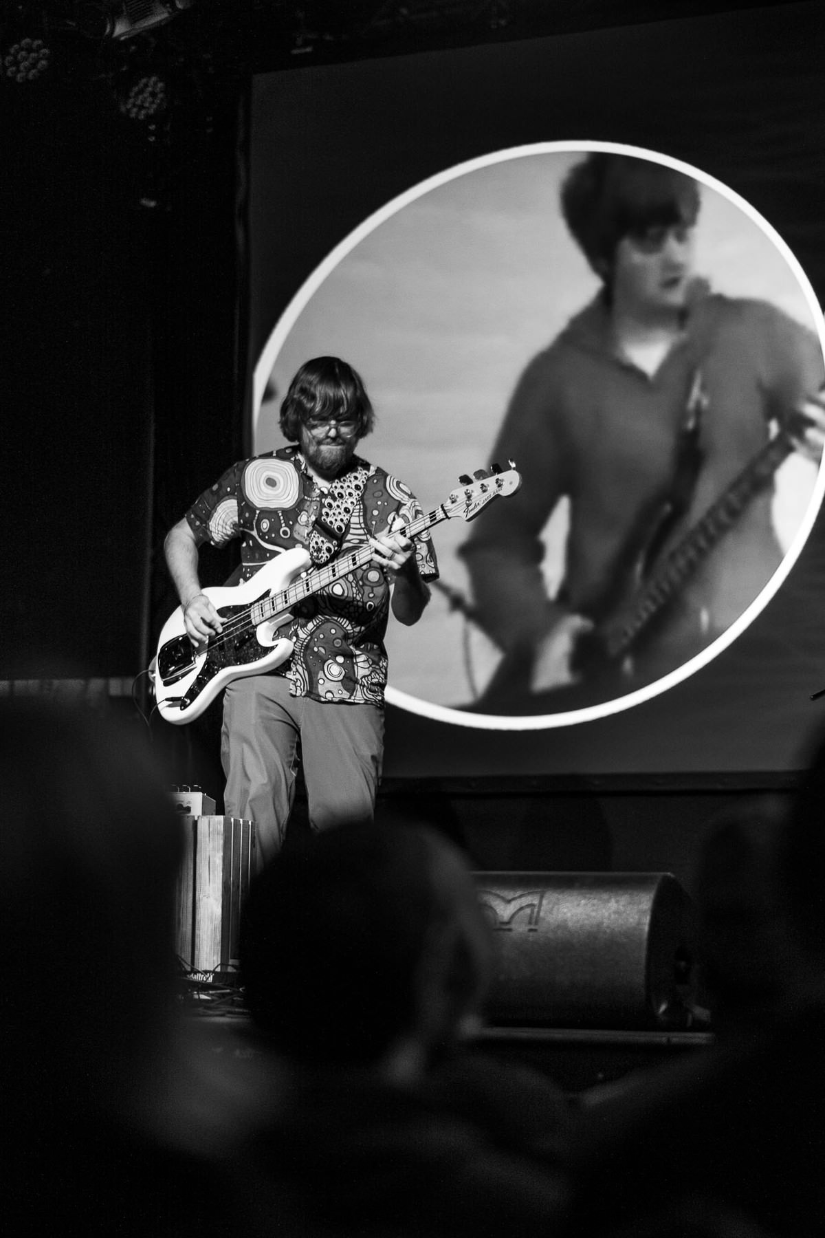 A black and white photo of Brad playing the bass. You can see the playing Brad on stage in front and a much younger brad playing bass on a picture behind him on the canvas.