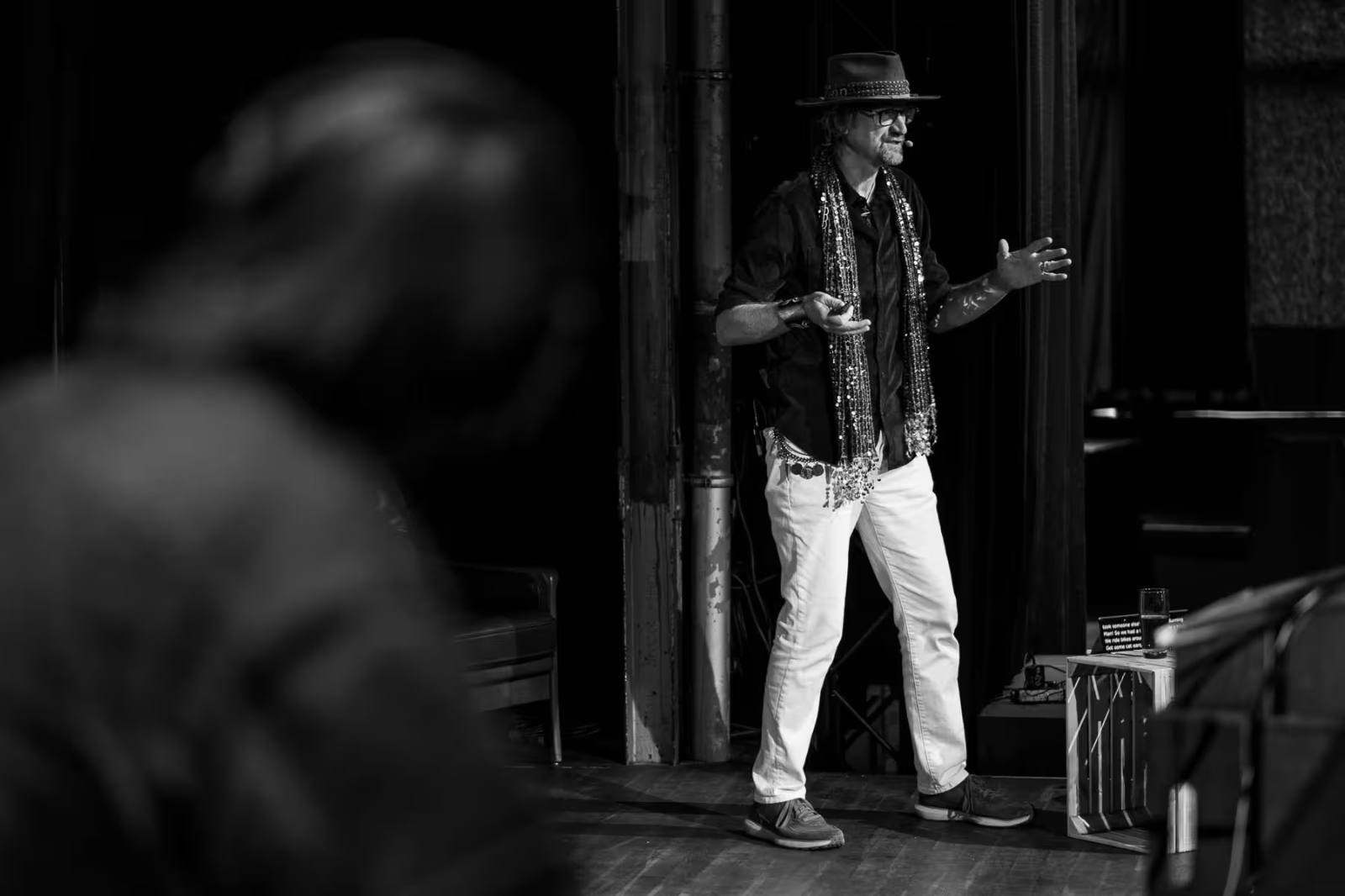 Black-and-white photo of Jared Ficklin speaking on stage at beyond tellerrand Berlin 2025. He wears a wide-brimmed hat, glasses, a dark shirt, and white jeans, with several long beaded necklaces draped around his neck. He gestures expressively with both hands while addressing the audience.