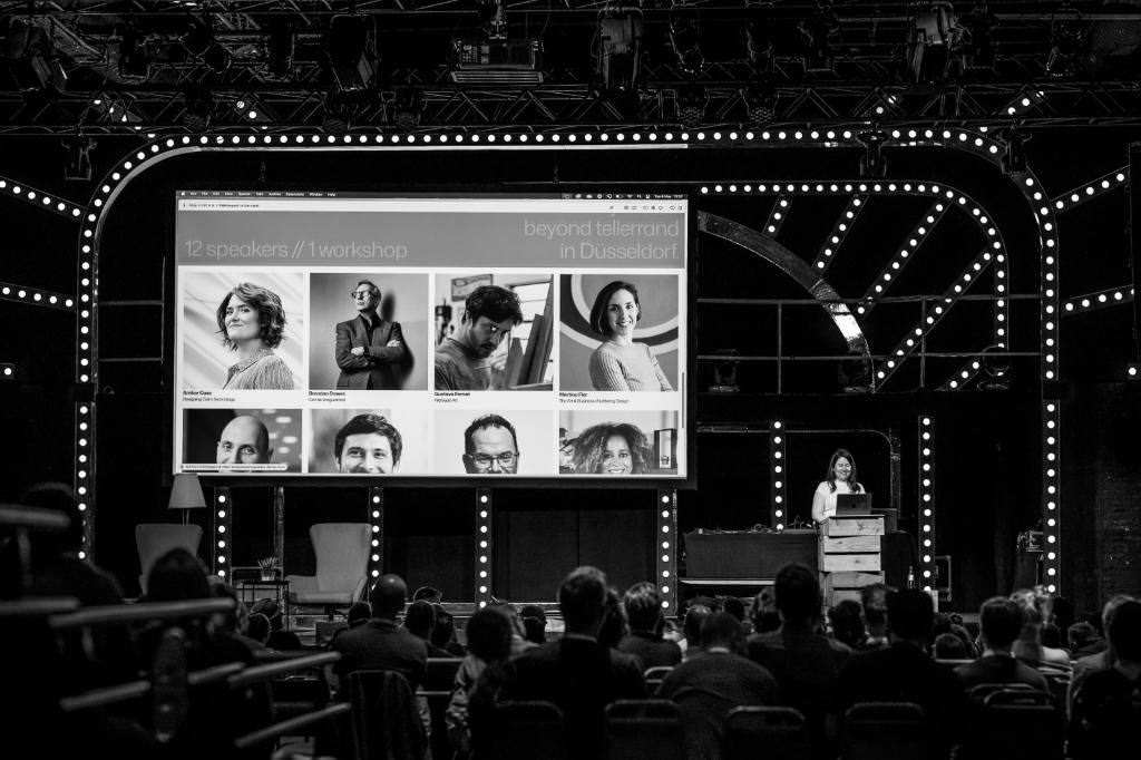 A black and white photo of Cyd Stumpel on stage at beyond tellerrand. The photo is taken from the back of the room and you can see the complete stage, surrounded by light bulbs. You can also see a full room on the lower half of the photo with attendees sitting in front of the stage, following the presentation.