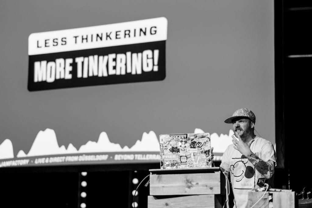 A black and white photo of Gavin Strange behind the speaker desk on stage at beyond tellerrand. He is gesturing with his hand and makes a grim face while explaining something that is important to him. On the slide, which you can only see parts of, in his back it says “Less Thinkering More Tinkering”.