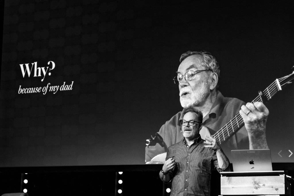 A black and white photo of Jason Pamental on stage at beyond tellerrand. You can see Jason from his belly upwards gesturing during his talk. In his back there is a large slide on a canvas with a photo of his dad. Text on the slide on the left states “Why? Because of my dad”.