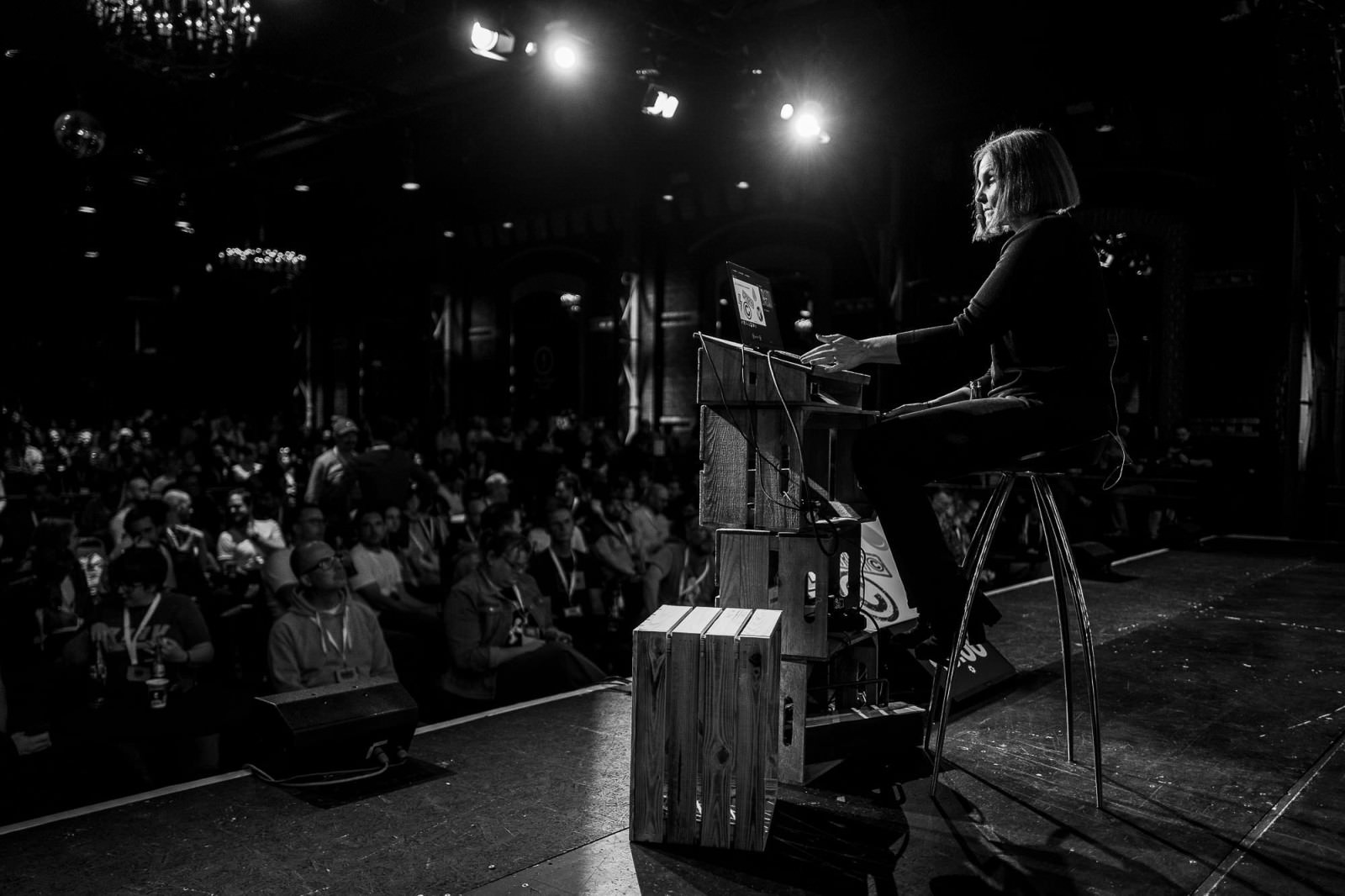 A black and white photo of Léonie Watson on stage at beyond tellerrand in Düsseldorf. The photo is taken from the side and you can see Léonie sitting behind the speaker desk on a high chair. The audience is on the left, blurred a little bit into the background.