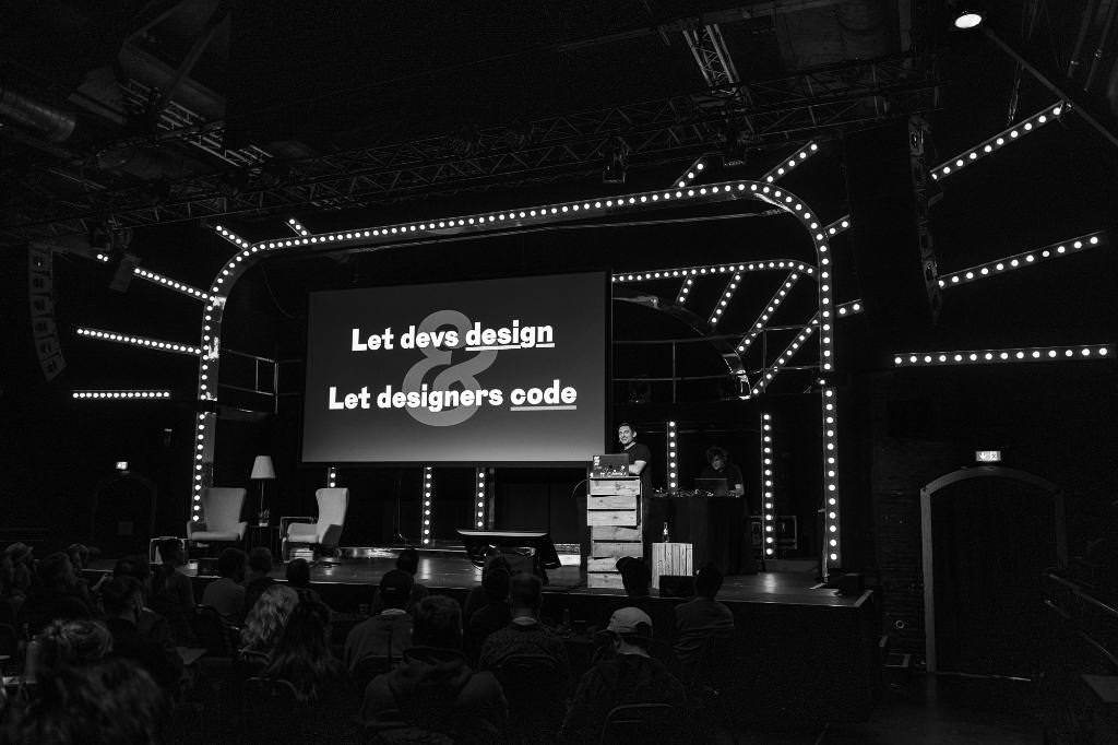 A black and white photo of Matthias on stage at beyond tellerrand in Düsseldorf. The slide in the back of him says “Let devs design. Let designer code.”.