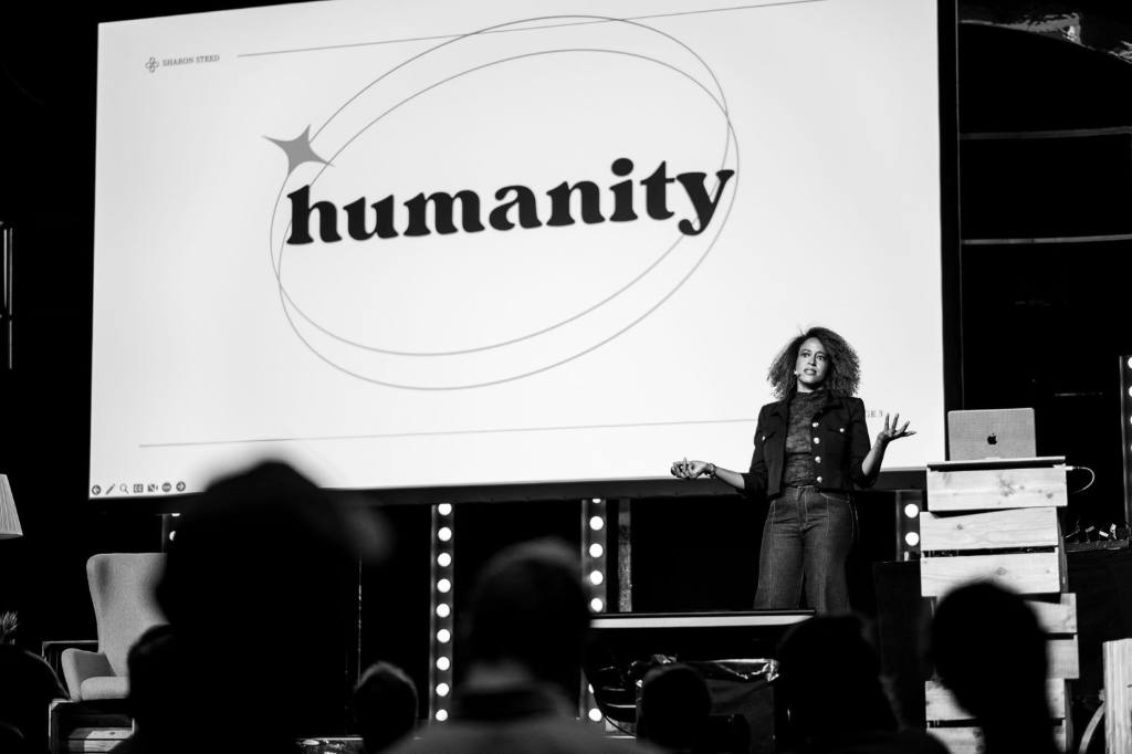 A black and white photo of Sharon Steed on stage at beyond tellerrand. You can recognise the audience in the foreground very blurry and out of focus. Sharon is on stage in focus and a slide in her back states in black type on white “humanity”. Two circles surround this word and a little star is overlapping the circles on the left.