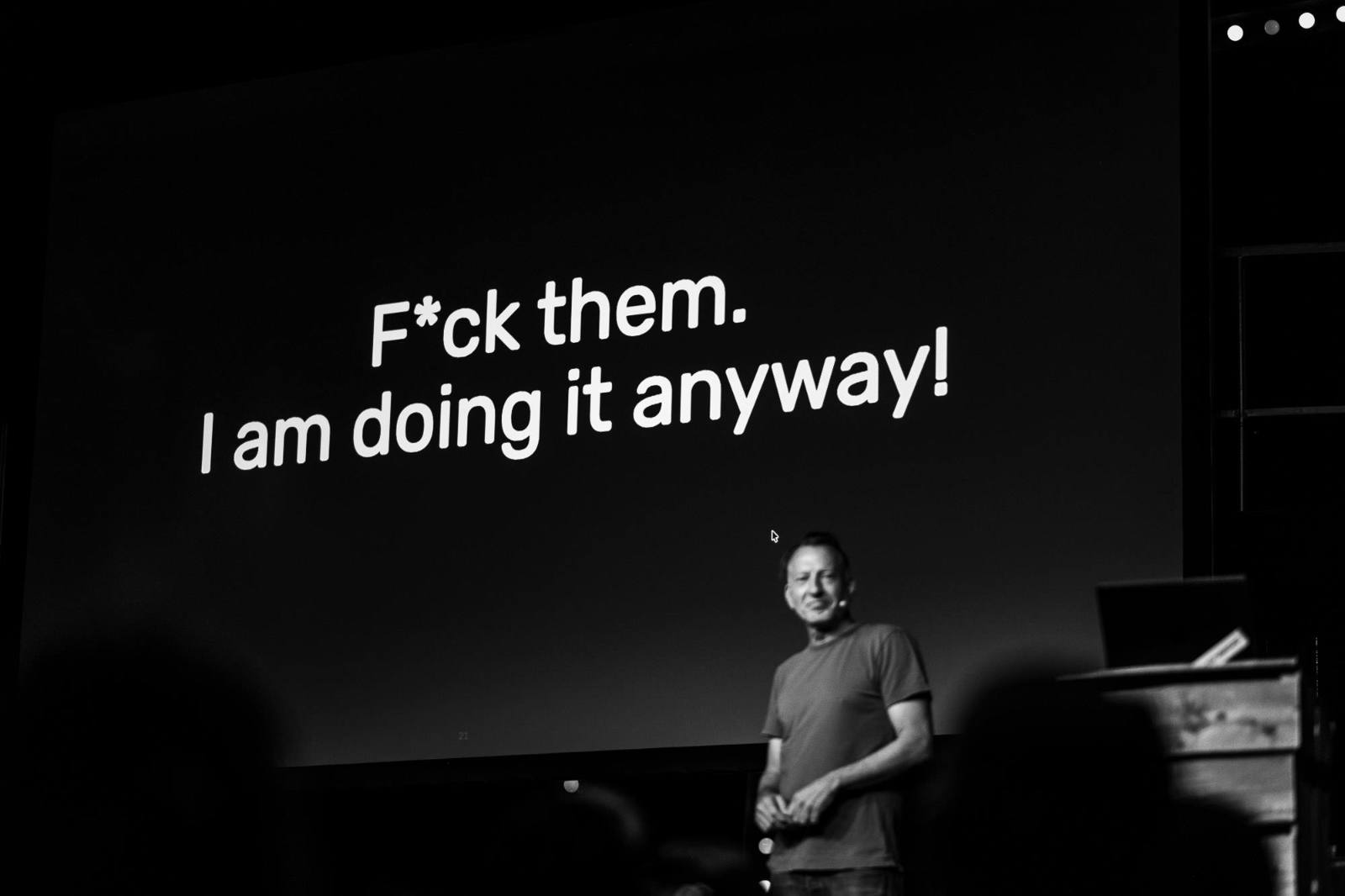 Black-and-white photo of Andr&eacute; Michelle standing on stage at beyond tellerrand, smiling slightly. Behind him, a large projection screen displays bold white text on a black background reading: &lsquo;F*ck them. I am doing it anyway!&rsquo;