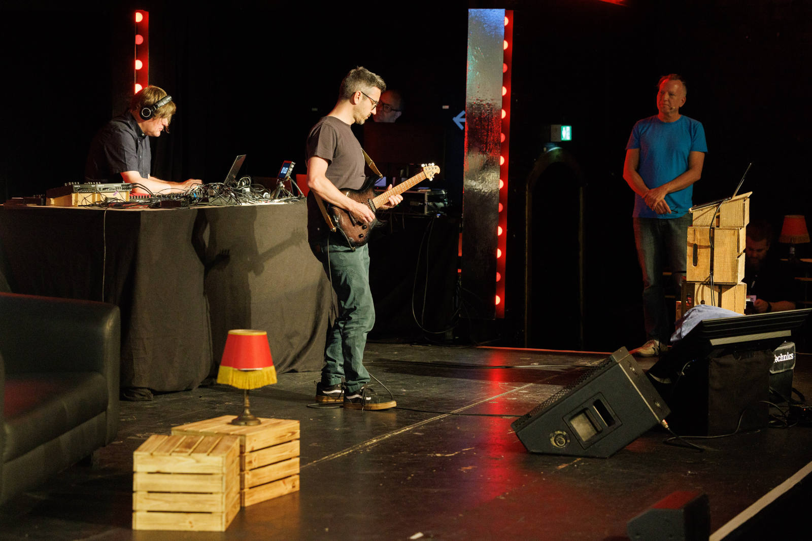 Stage view at beyond tellerrand showing Bastian Allgeier standing centre stage playing an electric guitar. On the right, Andr&eacute; Michelle in a blue t-shirt stands beside a stack of wooden crates. Wooden crates, a small red lamp, and a tipped-over stage monitor are scattered across the stage, framed by glowing red light columns.