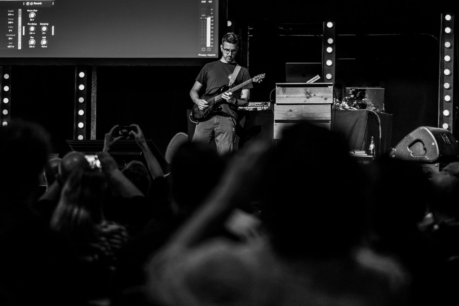Black-and-white photo of Bastian Allgeier playing electric guitar on stage at beyond tellerrand. Behind him, a large projection screen shows a reverb plugin interface. Silhouettes of audience members fill the foreground, with one person raising a phone to capture the moment.
