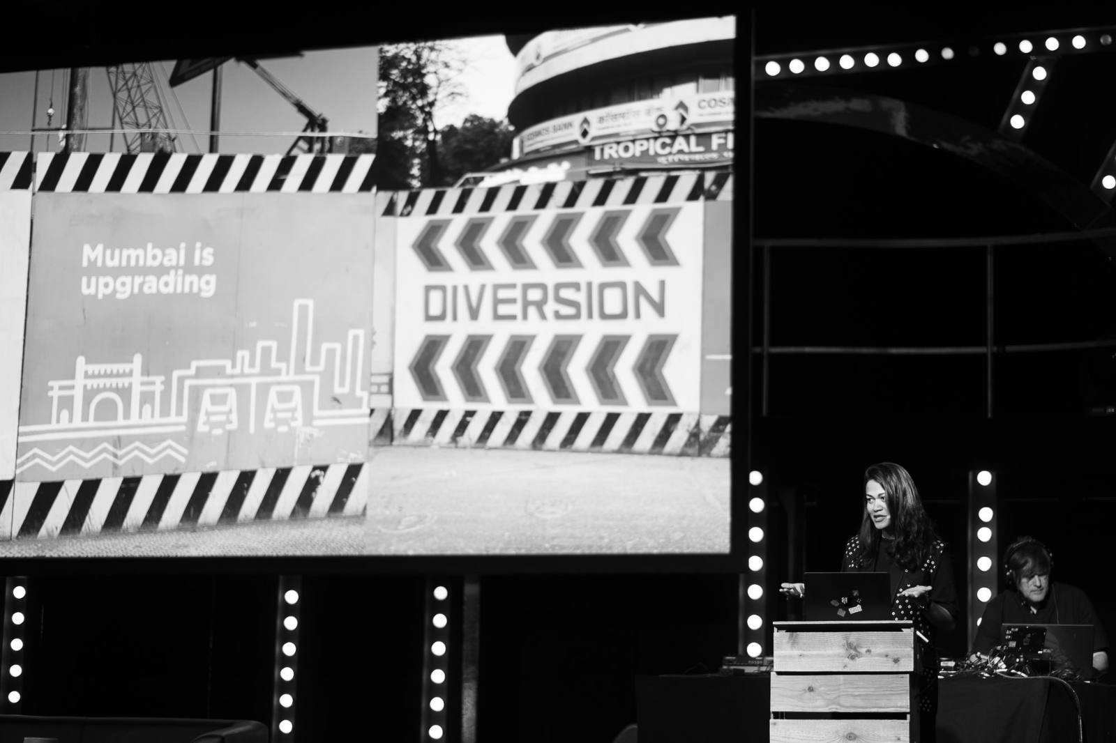 Black-and-white photo of Kimya Gandhi speaking on stage at beyond tellerrand D&uuml;sseldorf 2026. She stands behind a wooden lectern with a laptop, framed by rows of round stage lights. A technician sits to her right working at a console. Behind her, a large screen displays two photos from Mumbai: one showing a construction hoarding with a stylised city skyline and the text &lsquo;Mumbai is upgrading&rsquo;, the other showing a &lsquo;Diversion&rsquo; road sign with chevron arrows.