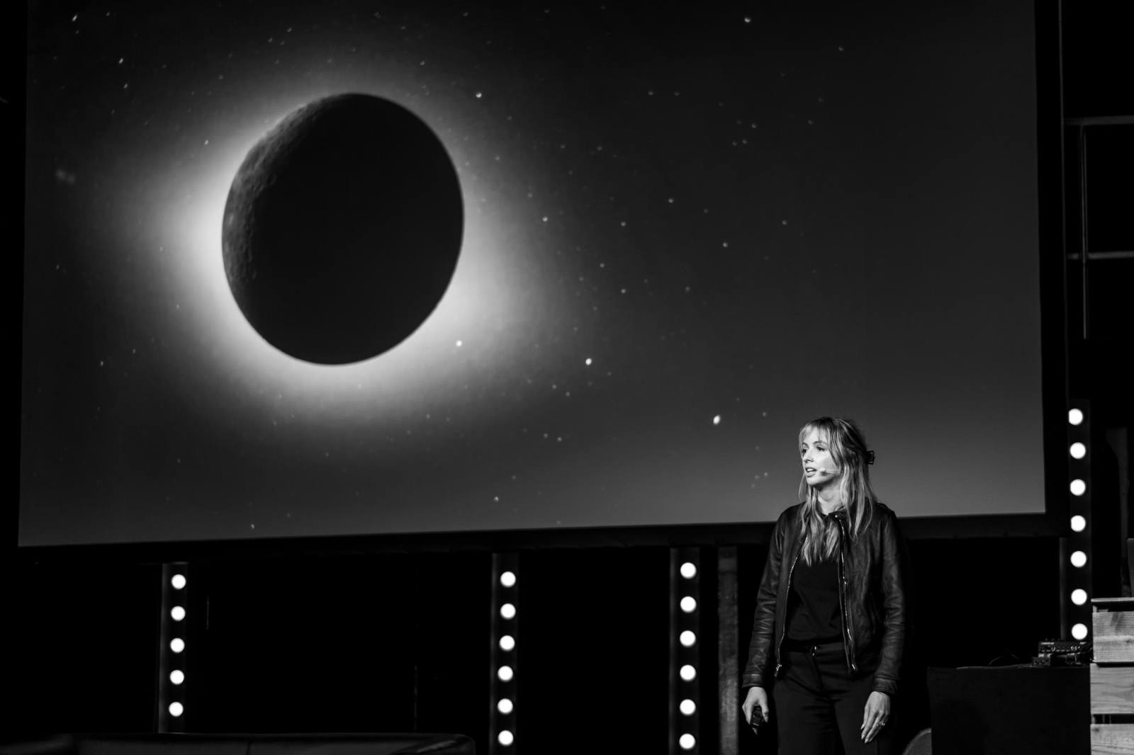 Black-and-white photo of Lauren Celenza speaking on stage at beyond tellerrand D&uuml;sseldorf 2026. She stands on the right wearing a leather jacket and headset microphone. A large screen behind her displays a striking image of a total solar eclipse, with the moon silhouetted against a glowing corona and starry sky. Illuminated stage light columns frame the scene.