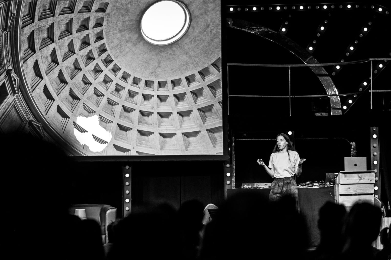 Black-and-white photo of Marjan van Aubel speaking on stage at beyond tellerrand Düsseldorf 2026. She wears a light t-shirt and gestures with both hands while addressing the audience. A large projection on the left shows the coffered dome and central oculus of the Pantheon in Rome. Stage lights and silhouettes of audience members frame the scene.