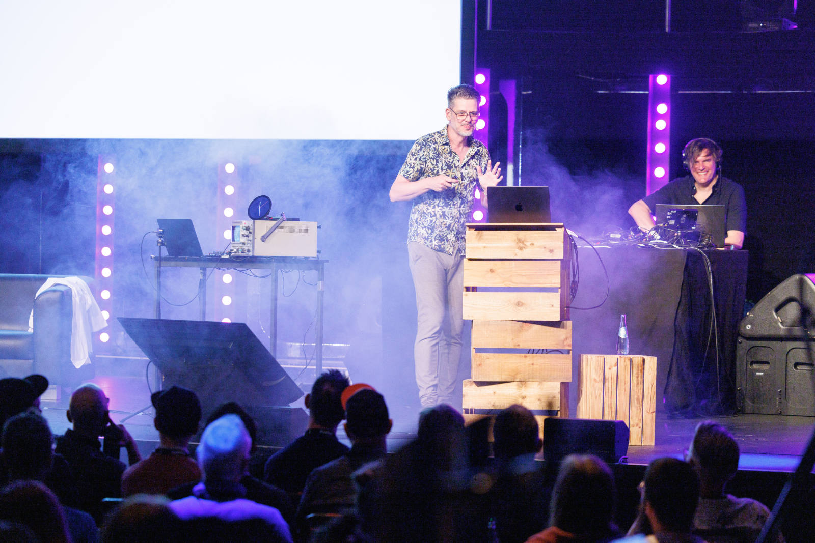 Niels Leenheer on stage at beyond tellerrand D&uuml;sseldorf 2026, smiling and gesturing as haze drifts across the stage, lit in blue and purple. He stands at a lectern made of stacked wooden crates with a laptop on top. Silhouettes of the audience fill the foreground.