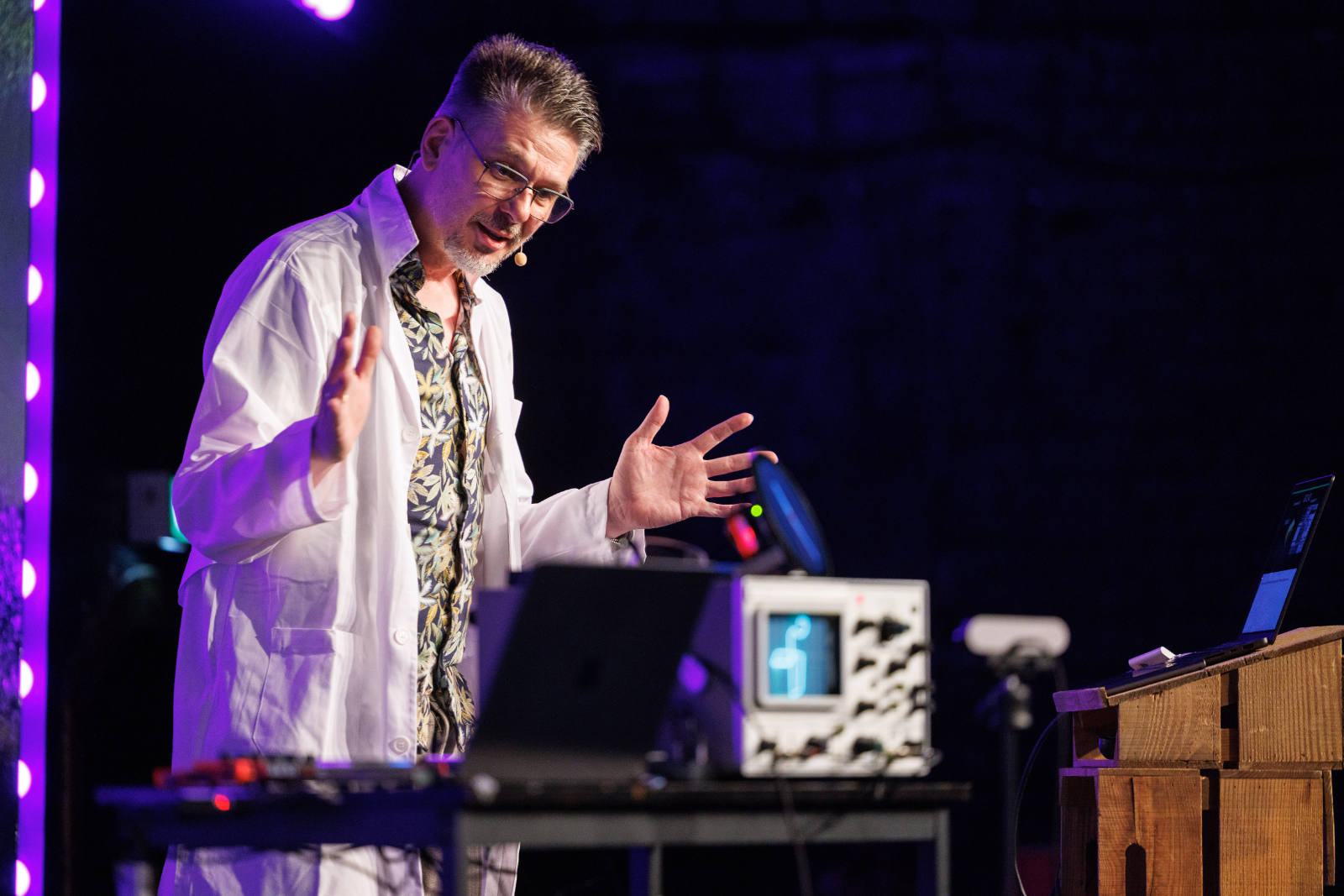 Niels Leenheer speaking on stage at beyond tellerrand D&uuml;sseldorf 2026, lit by purple stage lighting. He wears a white lab coat over a patterned shirt, glasses and a headset microphone, gesturing with both hands. In front of him sits a table with a laptop and a vintage oscilloscope displaying a glowing green graphic. A purple LED light column frames the left edge of the stage. link https://beyondtellerrand.com/photos/dusseldorf-2026-martin-kraft/mkr396058.jpg