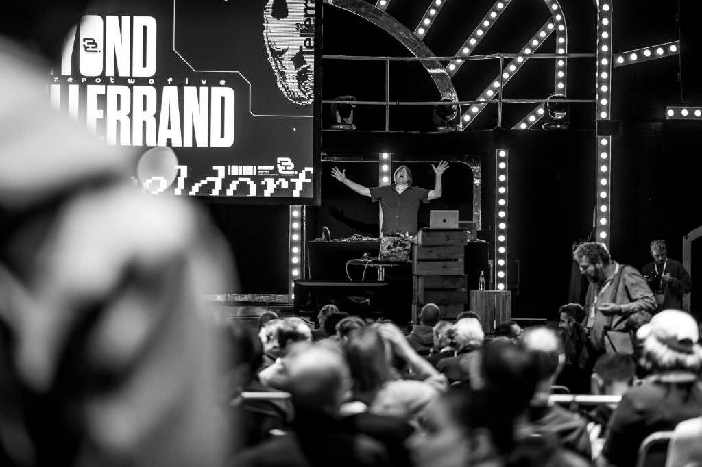 A photo from further back in the room. Chairs full with people. In the distance you can see the stage on which the DJ performs and seems to sing and dance.