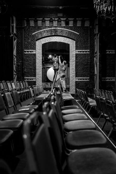 The empty theatre with a lot of empty chairs. In the back two people peeling of logo stickers that have been attached to the mirrors for the show.