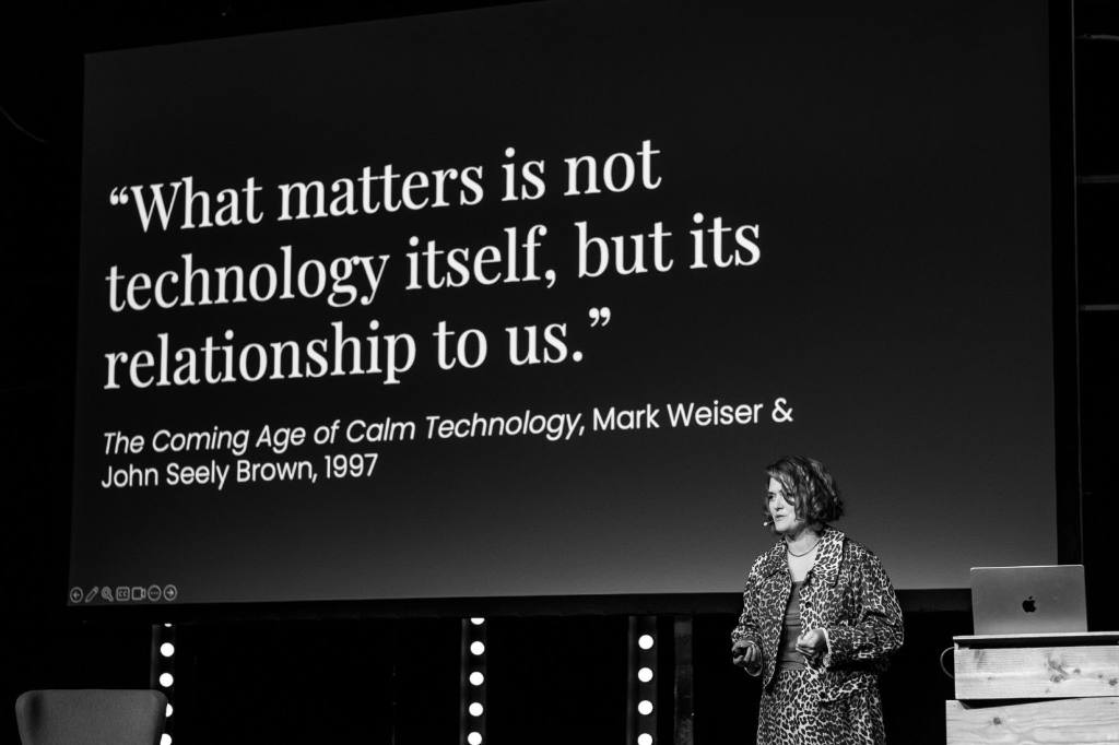 Amber Case during her talk. Amber wears a leopard dress and stands in front of the canvas with a slide with the quote “What matters is not technology itself, but its relationship to us.” and below the source “The Coming Age of Calm Technology, Mark Weiser &
John Seely Brown, 1997”.