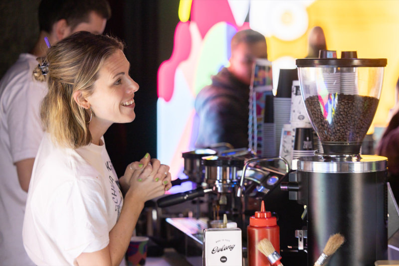The barista bar, organised and provided by our wonderful long time partners of sipgate. You can see a lady smiling on the left hand side, waiting for a person to order. On the right side of the photo, you can see the espresso machine and the grinder.