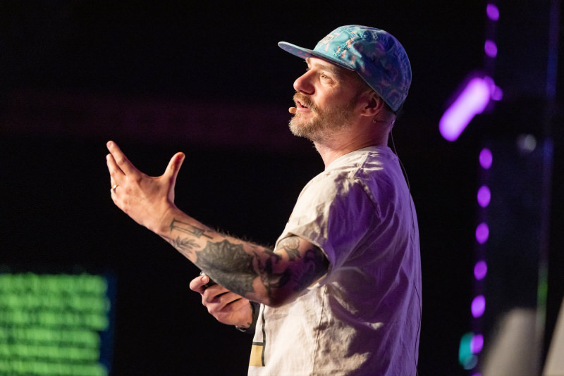 A close-up shot of Gavin Strange speaking on stage at beyond tellerrand. He performs a gesture that looks as if he is singing an aria.