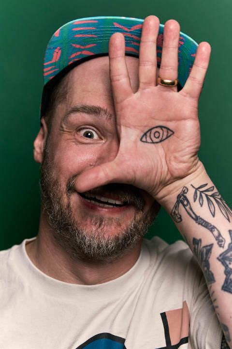 Portrait of Gavin Strange, holding hs left hand in front of his face in which he has a tattoo with an eye and it looks like the eye in his hand would be his actual eye. Shot in front of a green background.