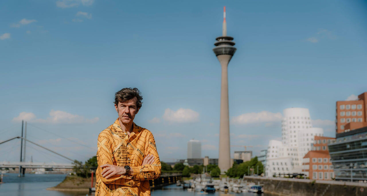 Stefan Sagmeister in front of the Düsseldorf skyline in which you can see the Fernsehturm and the Frank Gehry buildings at the Düsseldorf Rhine.