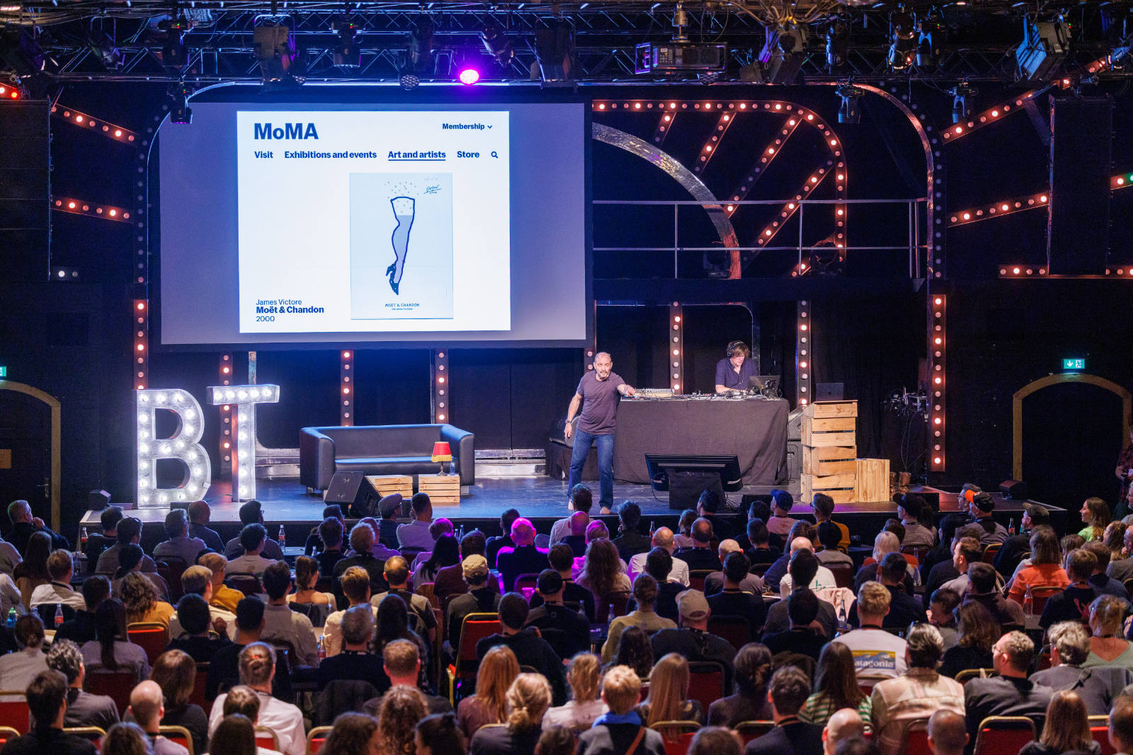 Wide shot of the beyond tellerrand stage during James Victore&rsquo;s talk. He stands barefoot at centre stage. Behind him, a large screen shows the MoMA website featuring his &lsquo;Mo&euml;t & Chandon, 2000&rsquo; poster. Illuminated &lsquo;BT&rsquo; marquee letters stand on the left of the stage, and the audience fills the foreground in silhouette.