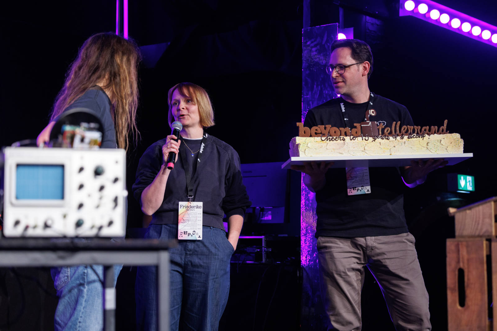 Three people on stage at beyond tellerrand. In the centre, Friederike Rennen speaks into a microphone. To the right, Lars Terlinden holds a large rectangular cake decorated with the &lsquo;beyond tellerrand&rsquo; logo and the text &lsquo;Cheers to 15 years of a btconf&rsquo;. On the left, Marc Thiele with long hair stands beside a vintage oscilloscope, partly turned away from the camera.