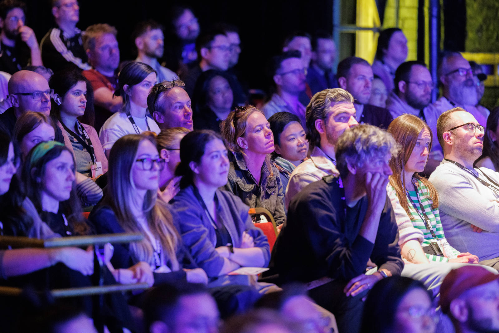 Close-up view of the beyond tellerrand audience seated in the venue, lit by warm purple and blue stage lighting. Dozens of attendees are visible in tight rows, attentively watching the stage, many wearing lanyards.