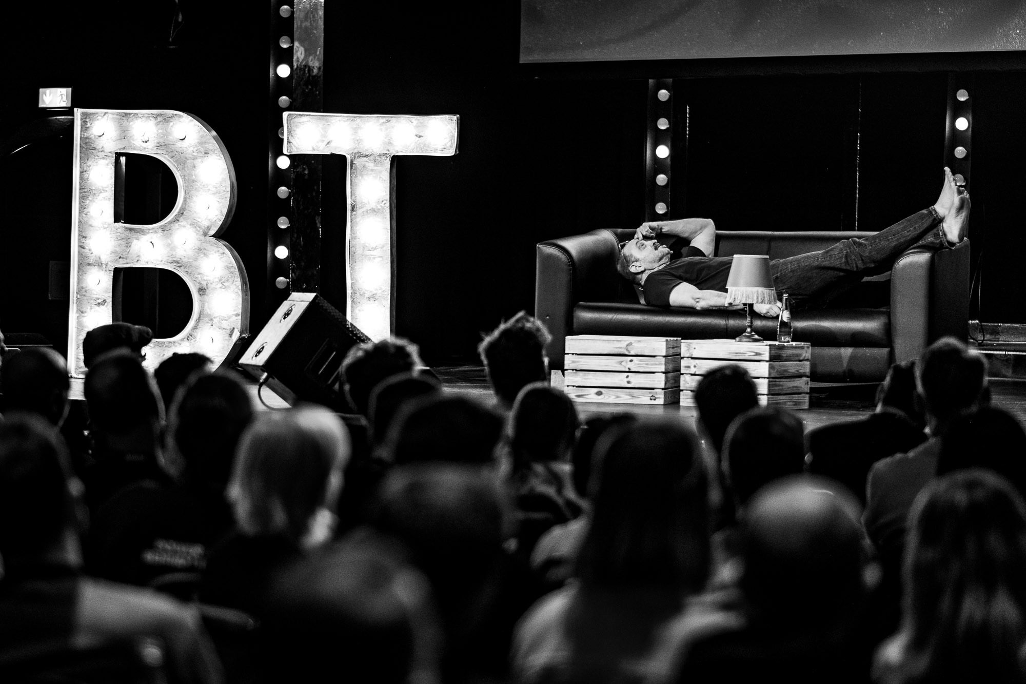 Black-and-white photo from beyond tellerrand D&uuml;sseldorf 2026: a barefoot speaker lies stretched out on a leather sofa on stage, one hand to his face mid-gesture, beside large illuminated "BT" marquee letters. The audience watches in silhouette in the foreground.