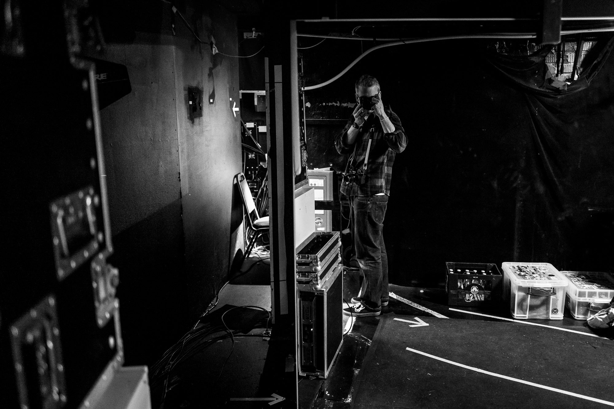 Black-and-white photo of Florian Ziegler standing backstage at beyond tellerrand D&uuml;sseldorf 2026, raising a camera to his eye. A second camera hangs from his neck. He's in a plaid shirt and jeans, surrounded by flight cases, cables, and crates of drinks in the dim wings of the venue.
