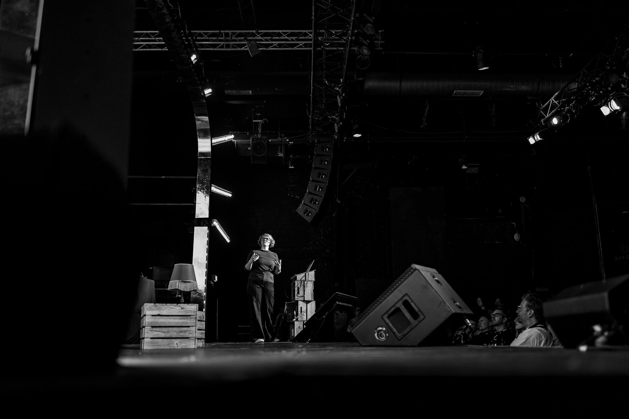 Black-and-white photo from beyond tellerrand D&uuml;sseldorf 2026: a speaker stands small and centred on a darkened stage, lit from above, mid-gesture with both hands raised. The venue's lighting rig and curved truss work loom around her, with a stage monitor and the silhouettes of a few audience members visible in the foreground.