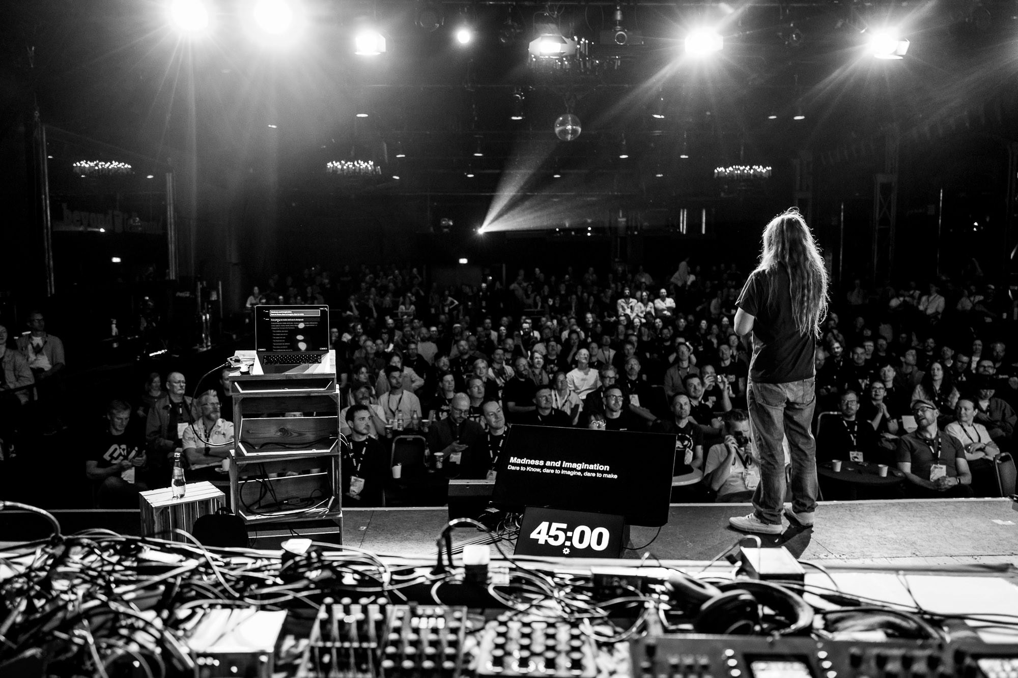 Black-and-white photo from behind the stage at beyond tellerrand D&uuml;sseldorf 2026: a long-haired speaker faces a full auditorium under bright stage lights, with a "45:00" timer and a monitor reading "Madness and Imagination" visible at their feet.