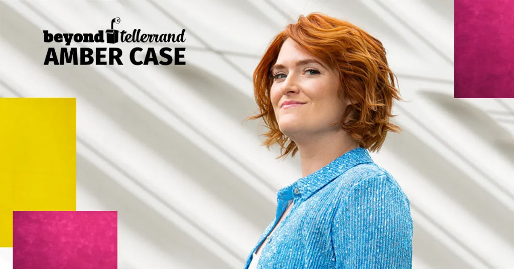 Portrait of Amber Case in which she wears a blue jacket and stands in front of a white background. She is smiling towards the camera and the photo is taken from an angle below her eye-line, so she is looking down.