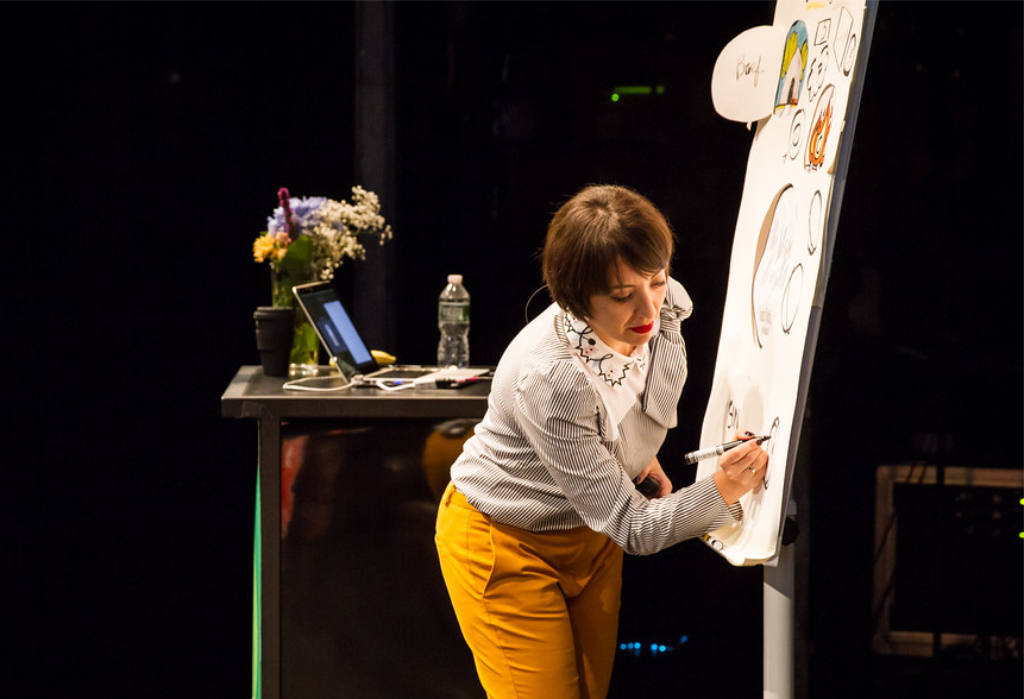 Chiara Aliotta on a dark stage, leaning towards a large flip chart and drawing on it with a black marker. She wears a striped blouse with an embroidered collar, mustard-yellow trousers and red lipstick. The flip chart is covered in hand-drawn illustrations and speech bubbles. Behind her stands a lectern with a laptop, a water bottle, a coffee cup and a small bouquet of flowers.