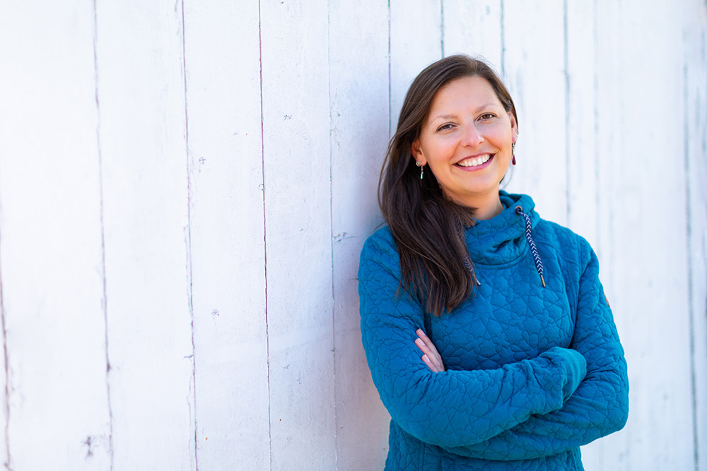 Portrait of Hannah Smith smiling at the camera, arms crossed, wearing a teal quilted hoodie. She leans against a white-painted wooden wall.