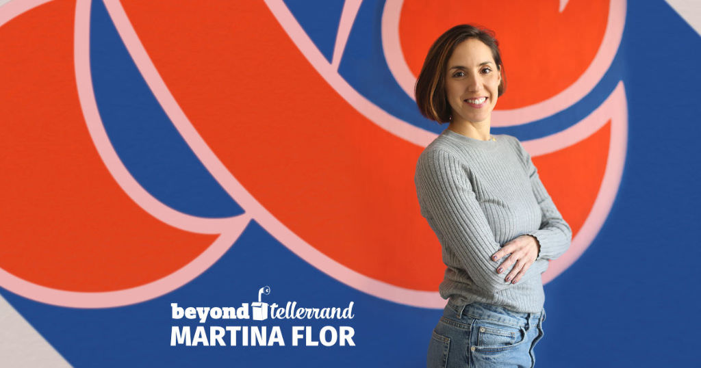 Martina Flor standing in front of a colourful wall with a blue, red and white graphic. She is turning the side to the viewer, crossing her arms, smilining into the camera.