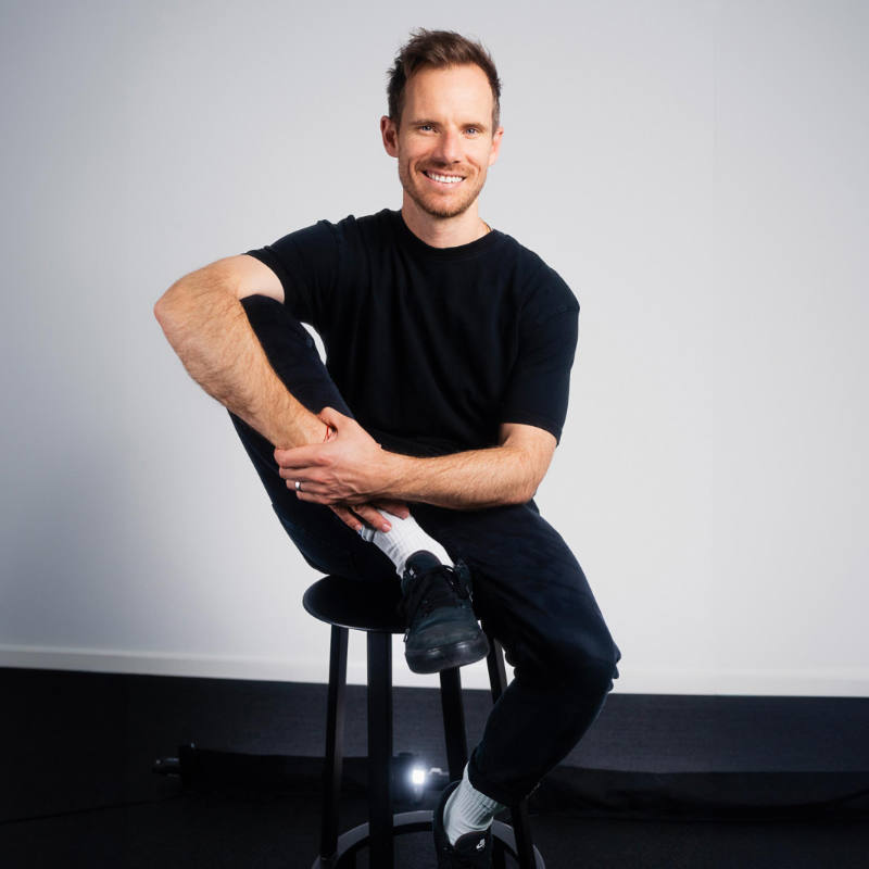 A photo of Andy Wyeth sitting on a bar stool, one leg up on the top of the chair, smiling into the camera.