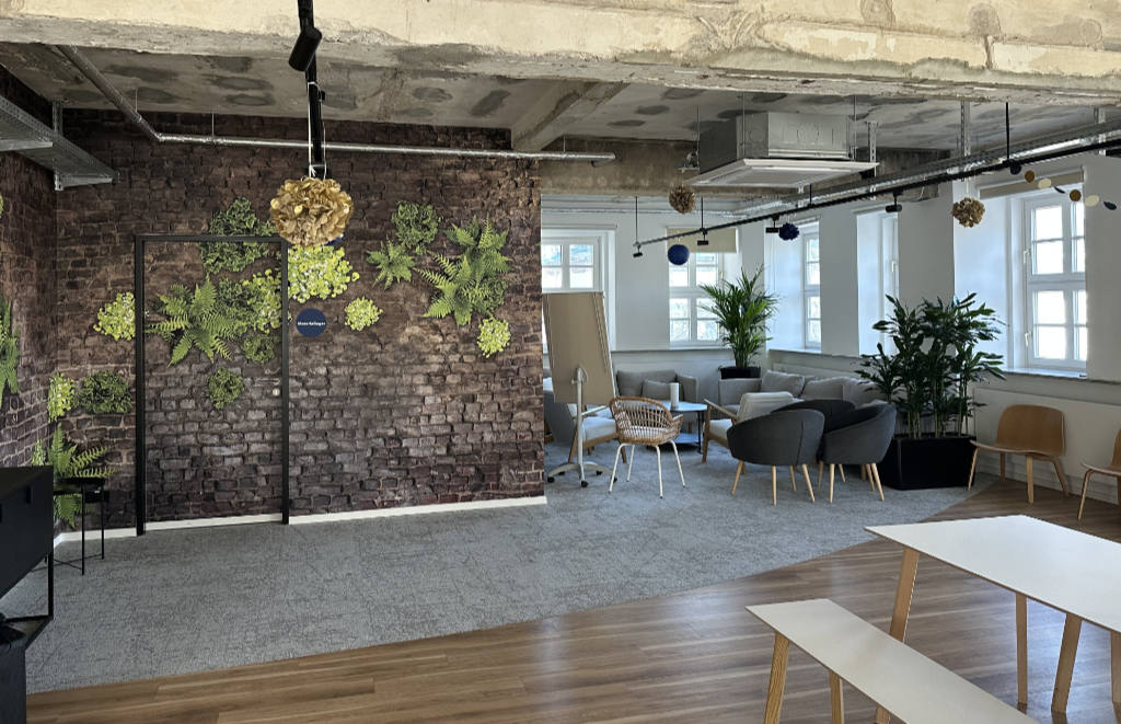 A photo of the meeting room at CHECK24. You can see a fake brick wall with plants growing out of those bricks on the left side of the photo and some cozy couches on the right side. The sun is shining through widows on the right side of the photo.