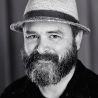 A portrait photo of Vasilis van Gemert. He has a longer beard, partly grey and black. On this photo he wears a head and smiles friendly into the camera..