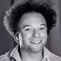 A portrait photo of Jude Pullen. He is a man, has curly wild hair and smiles into the camera.