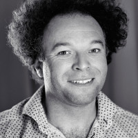 A portrait photo of Jude Pullen. He is a man, has curly wild hair and smiles into the camera.