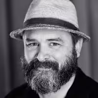 A portrait photo of Vasilis van Gemert. He has a longer beard, partly grey and black. On this photo he wears a head and smiles friendly into the camera..
